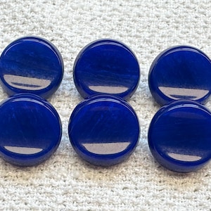 May include: Six round, polished, deep blue stones are arranged on a white, textured surface. The stones have a glossy finish and subtle marbling, reflecting light. The stones are likely decorative or used in jewelry.