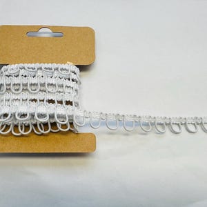 May include: A reel of white looped trim, suitable for sewing and craft projects. The trim is made of a soft, flexible material and is attached to a cardboard reel. The trim is a bright white colour.