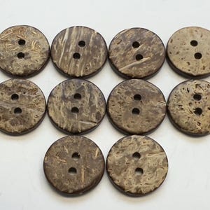 May include: A collection of ten round, natural-toned coconut shell buttons. Each button has two holes and a unique wood grain pattern, ranging from light tan to dark brown. These buttons are ideal for sewing and craft projects.