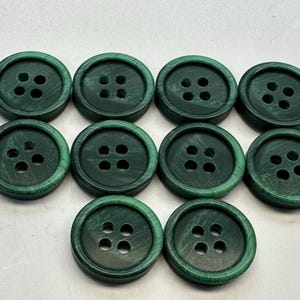 May include: A collection of ten dark green buttons with four holes each. The buttons are round with a slightly textured surface, and the color varies slightly across each button. The buttons are arranged in a scattered pattern.
