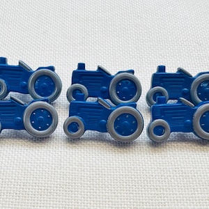 Puede incluir: Un conjunto de seis botones con forma de tractor azul. Cada botón presenta un cuerpo azul brillante con un volante gris y neumáticos grises. El diseño incluye líneas horizontales moldeadas en el lateral del cuerpo del tractor y detalles circulares.