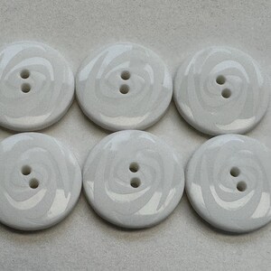 May include: Six white, round buttons with a swirled design. Each button has two holes for sewing. The buttons are a solid, opaque white with a subtle, swirling pattern in a lighter shade of white.
