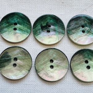 May include: Six round buttons with two holes each. The buttons have a pearlescent finish with shades of green, grey, and white. The buttons are arranged in two rows of three on a white fabric surface.