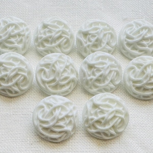 May include: Ten white, round buttons with a textured, woven design are displayed. The buttons are arranged on a white, textured surface, highlighting their intricate details and consistent look. These buttons are suitable for sewing and craft projects.