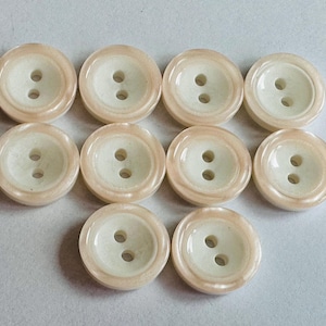 Pode incluir: Uma coleção de dez botões redondos. Cada botão tem um centro creme com dois furos e um anel externo de cor pêssego claro. Os botões estão dispostos em um padrão ligeiramente curvo.