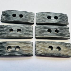 May include: Six rectangular buttons with two holes each. The buttons have a grey and white wood grain pattern. The buttons are slightly curved and have rounded edges. These buttons are suitable for sewing projects.