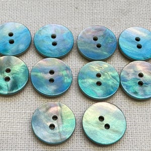May include: Ten round buttons with a pearlescent finish in shades of blue and green. Each button has two holes for sewing. The buttons are arranged on a neutral, textured surface.