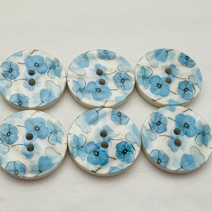May include: Six round, white, wooden buttons with a blue floral pattern. The buttons have two holes each.