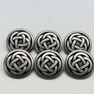 Könnte beinhalten: Sechs silberfarbene Knöpfe mit einem keltischen Knotenmuster. Die Knöpfe sind rund mit einem erhabenen, detaillierten Knotenmuster in der Mitte. Sie sind in zwei Reihen zu je drei Stück angeordnet.