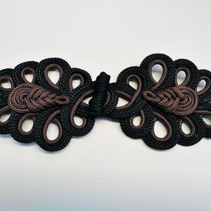 Peut inclure: Fermeture de manteau décorative noire et marron. La conception symétrique est faite de cordon tressé, avec un nœud central et des extrémités en boucle. Le schéma de couleurs est une combinaison de noir et de marron.