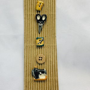 Puede incluir: Una colección de botones con temática de costura, exhibidos en una tira de cartón corrugado. Los botones incluyen un botón amarillo, tijeras, una bobina de hilo, un botón de madera y una máquina de coser. Los botones son de varios colores, incluyendo amarillo, negro y marrón.