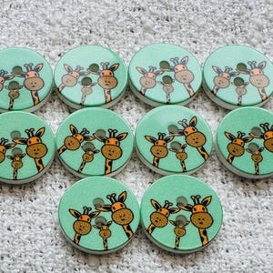 May include: Ten round, wooden buttons with a light teal background and a giraffe family design. The giraffes are brown and tan with black accents. Each button has two holes for sewing.