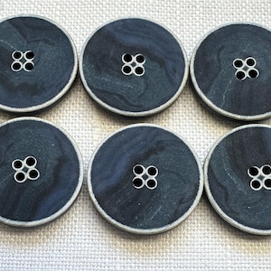 May include: Six round, dark blue buttons with a marbled effect and a silver-colored rim. Each button has four holes arranged in a square pattern. The buttons are displayed on a light-colored fabric.