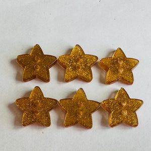 May include: Six gold glitter star-shaped buttons. Each button has two holes for sewing. The buttons are arranged in two rows of three on a white background. These buttons are suitable for various craft projects.