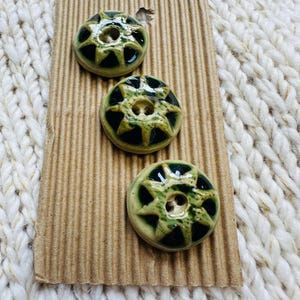 May include: Three round ceramic buttons arranged vertically on a rectangular piece of corrugated cardboard. Each button features a glossy finish with a dark green centre and a lighter, mottled green star-shaped pattern radiating outward.