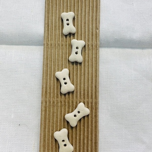 May include: Five off-white, bone-shaped buttons with two central holes each, arranged vertically on a rectangular piece of brown, vertically-ribbed corrugated cardboard that features a small circular hole at the top center for hanging.