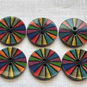 May include: Six circular buttons with a vibrant, radial pattern. Each button has a black centre with two holes and a design of radiating lines in red, orange, yellow, green, blue, and purple. The buttons are approximately 3.8 cm in diameter.