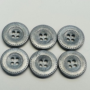 Könnte beinhalten: Sechs runde, silberfarbene Knöpfe mit strukturiertem, metallischem Finish. Jeder Knopf hat vier Löcher und einen dekorativen, gepunkteten Rand. Die Knöpfe sind in zwei Dreierreihen angeordnet.