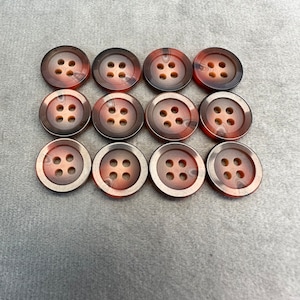 Puede incluir: Doce botones marrones y beige con cuatro agujeros cada uno. Los botones tienen un patrón moteado.
