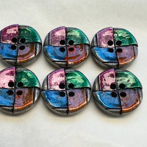 May include: A set of six round, four-hole buttons arranged in two rows. Each button features a segmented design with metallic, glossy sections in shades of pink, green, blue, and copper, separated by dark, recessed lines across the surface.