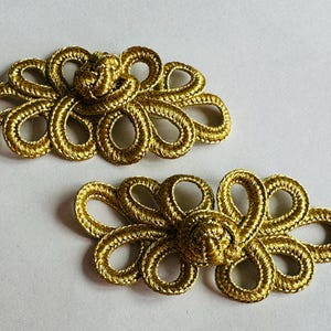 May include: Two gold-coloured, ornate floral clasps. Each clasp features a central knot surrounded by looped, petal-like designs. The clasps are made of a shiny, woven material, and are set against a white background.