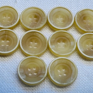 May include: Ten round, light yellow buttons with four holes each are arranged on a white textured surface. The buttons have a slightly translucent appearance and a raised rim. The buttons are a classic sewing or craft supply.