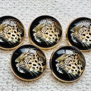 Puede incluir: Cinco botones redondos con bordes dorados, cada uno con el perfil de un leopardo sobre un fondo negro. El leopardo tiene pelaje marrón con manchas negras. Los botones están dispuestos en un patrón circular.