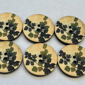 May include: Six round wooden buttons with a botanical design. Each button features a light beige background with a dark green and olive green leaf and vine pattern. The buttons have two holes for sewing.