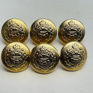 Puede incluir: Seis botones de color dorado con un diseño en relieve que presenta una corona y un símbolo. Los botones son redondos y tienen una superficie lisa.