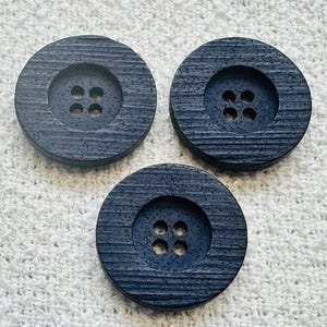 May include: Three circular dark navy blue buttons with a textured, wood-grain appearance. Each button features a recessed center with four holes arranged in a square pattern. The surface of each button has horizontal ridges and a speckled finish.