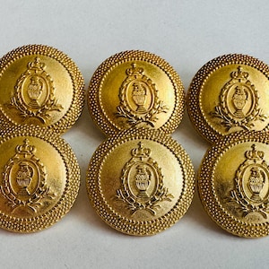 Könnte beinhalten: Sechs goldfarbene Knöpfe mit detailliertem Design. Jeder Knopf zeigt ein zentrales Emblem einer Krone über einer Vase, umgeben von einem Lorbeerkranz. Die Knöpfe haben einen strukturierten, perlenbesetzten Rand.