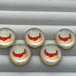 May include: Five round, gold-rimmed, glass buttons with a white background and a picture of a strawberry shortcake dessert. The buttons have the text "Frost Tea" printed on them.