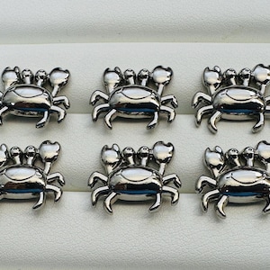 May include: A set of six small, polished silver-tone metal crab charms. Each crab features a rounded body, two raised eyes, and two prominent claws extended upwards. The metallic surface is smooth and reflective, showing a consistent design.