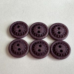 May include: Six dark purple buttons with a speckled texture and a decorative outer edge. Each button has two holes for sewing. The buttons are arranged in two rows of three on a white background.