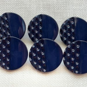 Könnte beinhalten: Ein Set aus sechs kreisförmigen marineblauen Knöpfen, angeordnet in zwei Reihen zu je drei Stück. Jeder Knopf hat ein geteiltes Design, wobei eine Hälfte ein strukturiertes Muster aus kleinen ovalen Vertiefungen und die andere eine glatte Oberfläche aufweist.