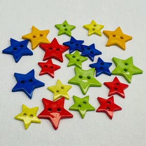 May include: A collection of star-shaped buttons in various colors, including yellow, red, blue, and green. Each button has two holes for sewing. The buttons are scattered on a white surface.