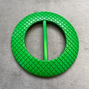 Puede incluir: Una hebilla de cinturón de plástico verde con un patrón de diamantes. La hebilla tiene una barra de metal que atraviesa el centro.