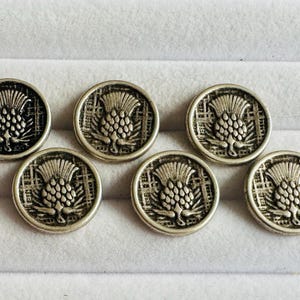 May include: Seven silver-toned buttons with a detailed thistle design. The buttons are round with a raised thistle motif in the center, surrounded by a textured border. The buttons are arranged on a white surface.