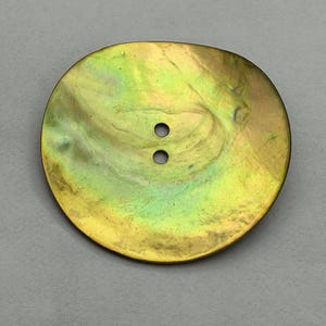 Może przedstawiać: Duży, iryzujący guzik o perłowym połysku. Guzik ma owalny kształt i dwa małe otwory. Kolory wahają się od złotego do zielonego, z subtelnymi wirami i wzorami.