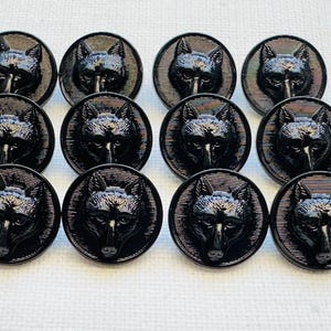 May include: A set of twelve black buttons, each with a wolf head motif. The buttons are circular, with a detailed wolf face at the centre and a textured edge. The buttons are made of a dark, shiny material.