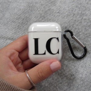 May include: White Airpods case with a black 'LC' monogram and a black carabiner clip.