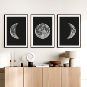 Op de afbeelding: Drie zwart-witte ingelijste prints van de maan in verschillende fasen. De prints hangen aan een witte muur boven een houten kast met een lamp, kaarsen en decoratieve objecten.