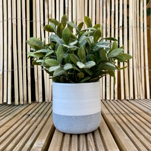 May include: A white ceramic planter with a grey speckled base, filled with a green artificial plant. The planter is sitting on a wooden deck.
