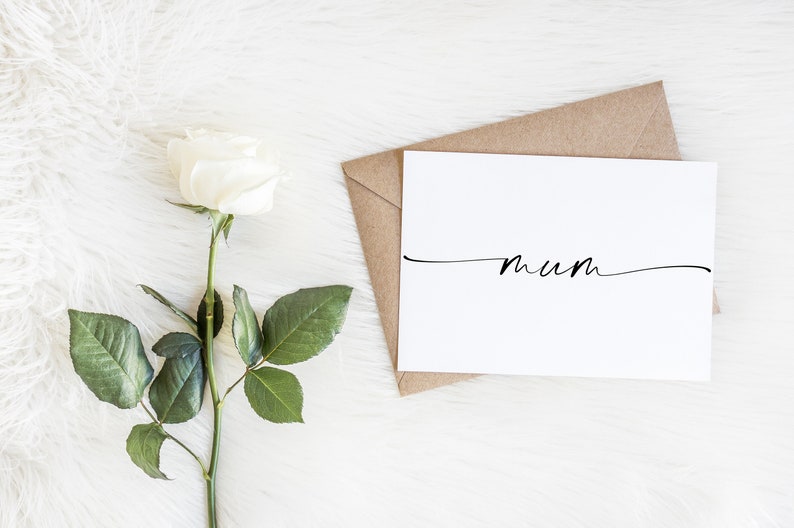 May include: A white card with the word "mum" written in cursive on it. The card is sitting on a brown envelope. A single white rose with green leaves is on the left side of the image.