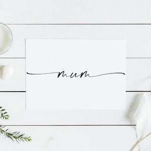 May include: A white card with the word "mum" written in black cursive script on a white wooden background. There are decorative items around the card, including a candle, a seashell, and a sprig of greenery.
