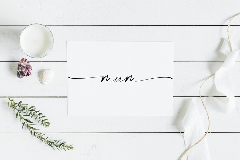 May include: A white card with the word "mum" written in black cursive script on a white wooden background. There are decorative items around the card, including a candle, a seashell, and a sprig of greenery.