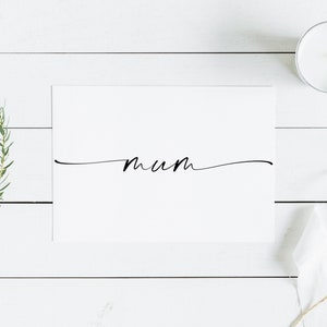 May include: A white card with the word "mum" written in black cursive script on a white wooden background. There is a small white candle and a piece of white fabric with twine on the right side of the image.