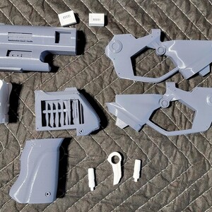 Peut inclure: Ensemble de pièces de pistolet grises imprimées en 3D, comprenant un canon avec un embout orange, une poignée et divers autres composants. Plusieurs petits rectangles blancs sont également présents. Les pièces sont disposées sur une surface sombre et texturée.