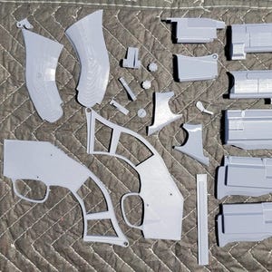 May include: A set of grey plastic parts for a model gun. The parts are laid out on a grey patterned surface.