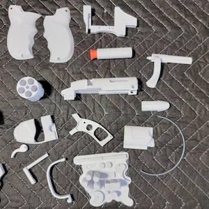 May include: A set of white plastic parts for a toy gun. The parts include a barrel, a handle, a trigger, and a magazine. The parts are laid out on a grey fabric surface.